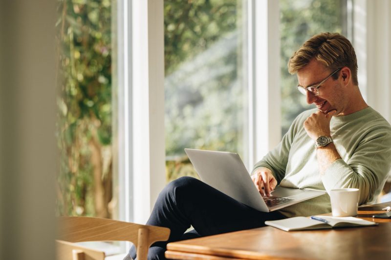 Young man using laptop and smiling at home. Man sitting by table working on laptop computer.