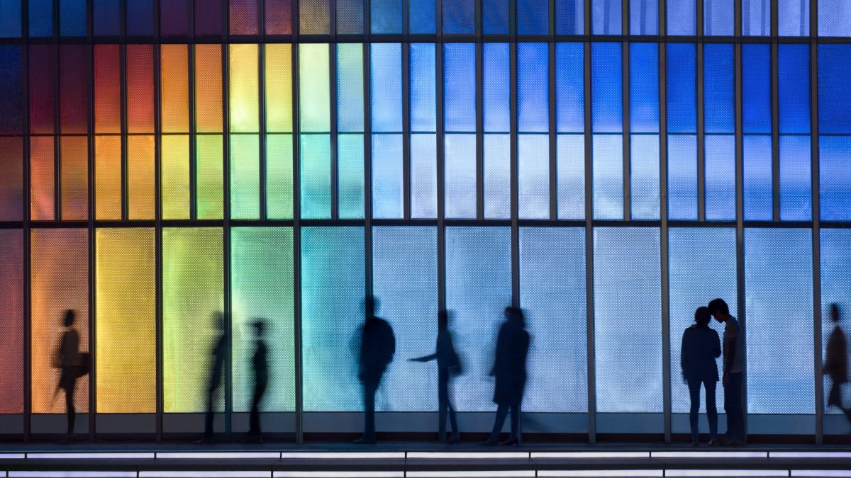 Silhouettes of people passing LED illuminated facade with continuously changing colors.