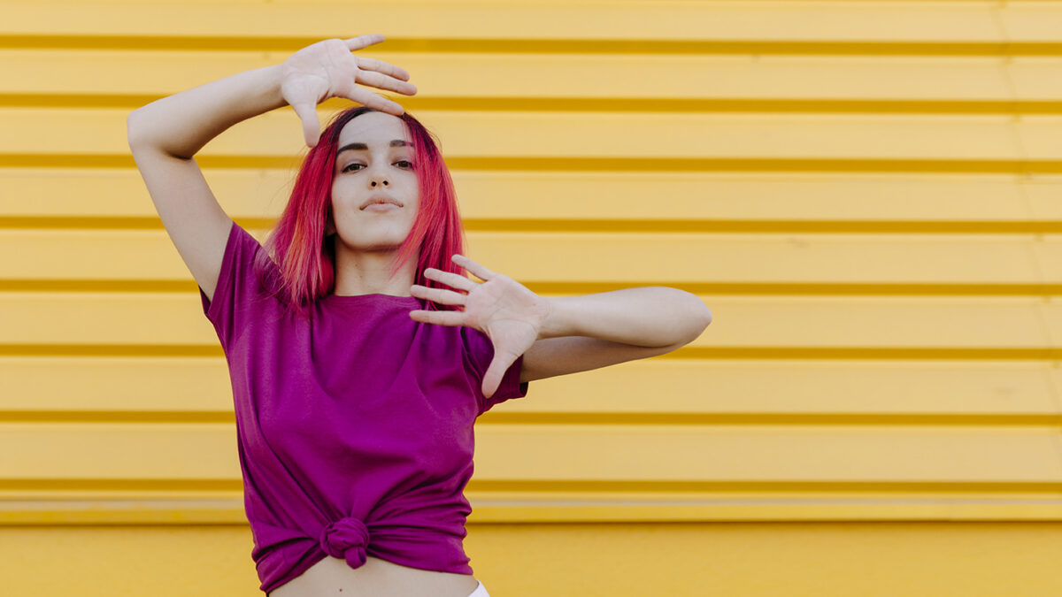 Confident young woman with dyed hair dancing against yellow wall