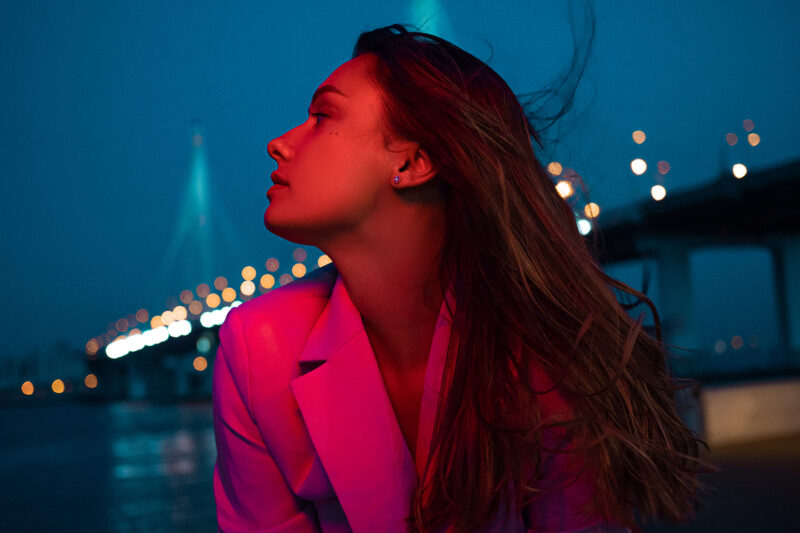 young stylish brunette in lights of night city, neon lighting, red light on her face. young woman on an evening walk around city, cable-stayed bridge and panorama of the night city on background