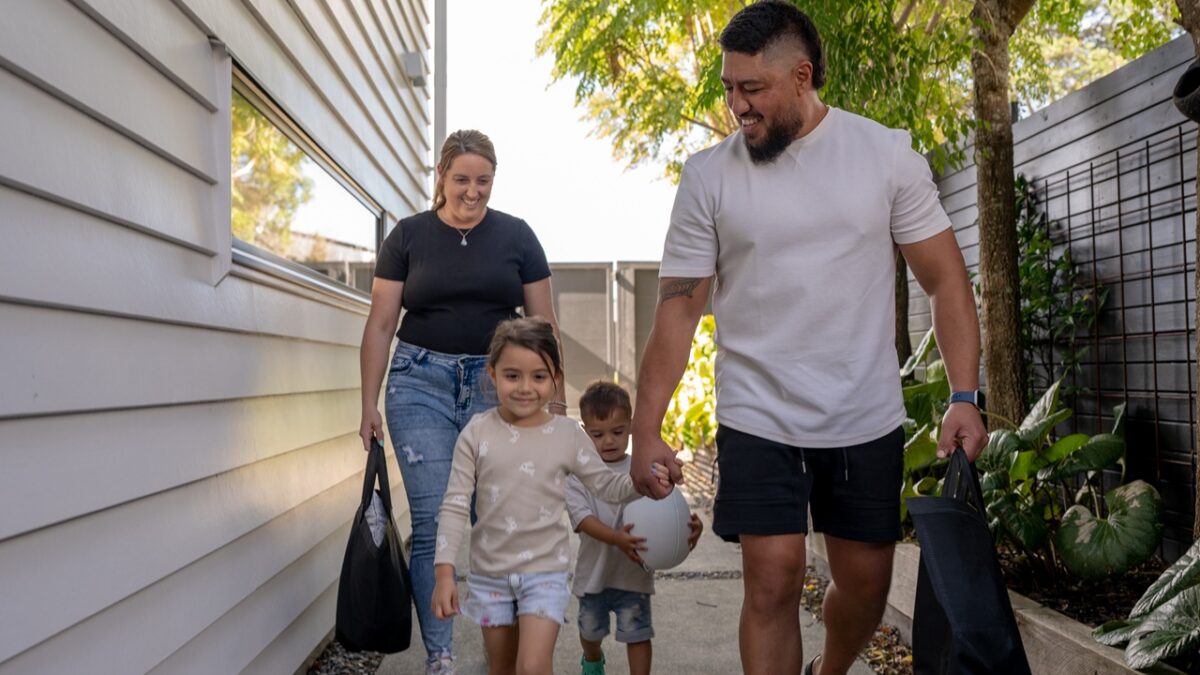 A cheerful family of four walks down a garden path beside their home. The parents carry reusable bags while their children walk ahead